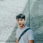 Casual portrait of a young Asian man posing against a textured stone wall, in outdoor daylight.