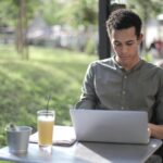 Young man working on a laptop outside in a casual and relaxed setting, enjoying a drink.