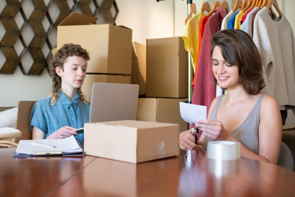Two women organizing and managing an online clothing store with boxes and a laptop.